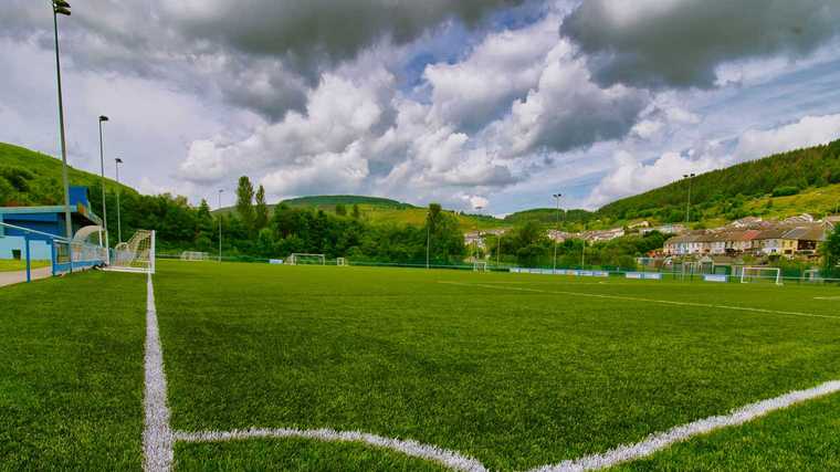 Natural grass vs artificial turf comparison showing a green athletic field surrounded by hills and cloudy skies.