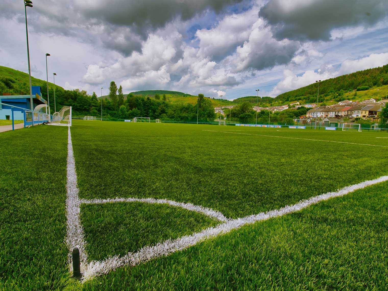Natural grass vs artificial turf comparison showing a green athletic field surrounded by hills and cloudy skies.