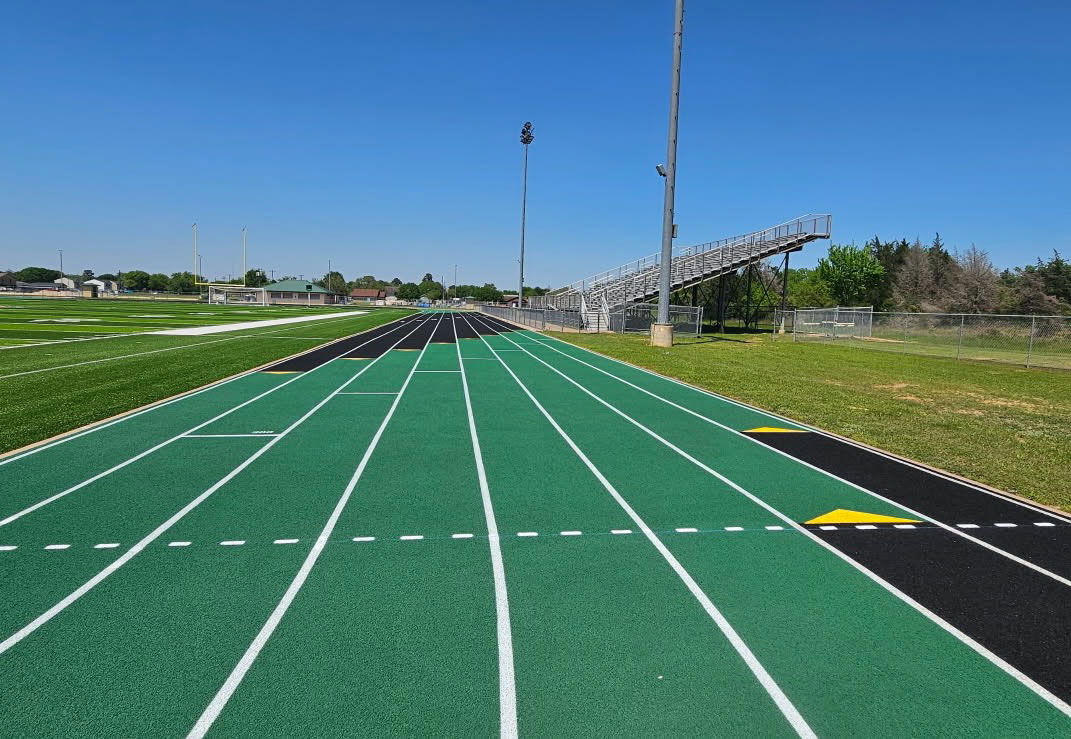 Ssc-values-surfaces-2560-1774 - Symmetry Sports Construction Green running track with white lane lines extending toward metal bleachers under a clear blue sky, on a sunny day.