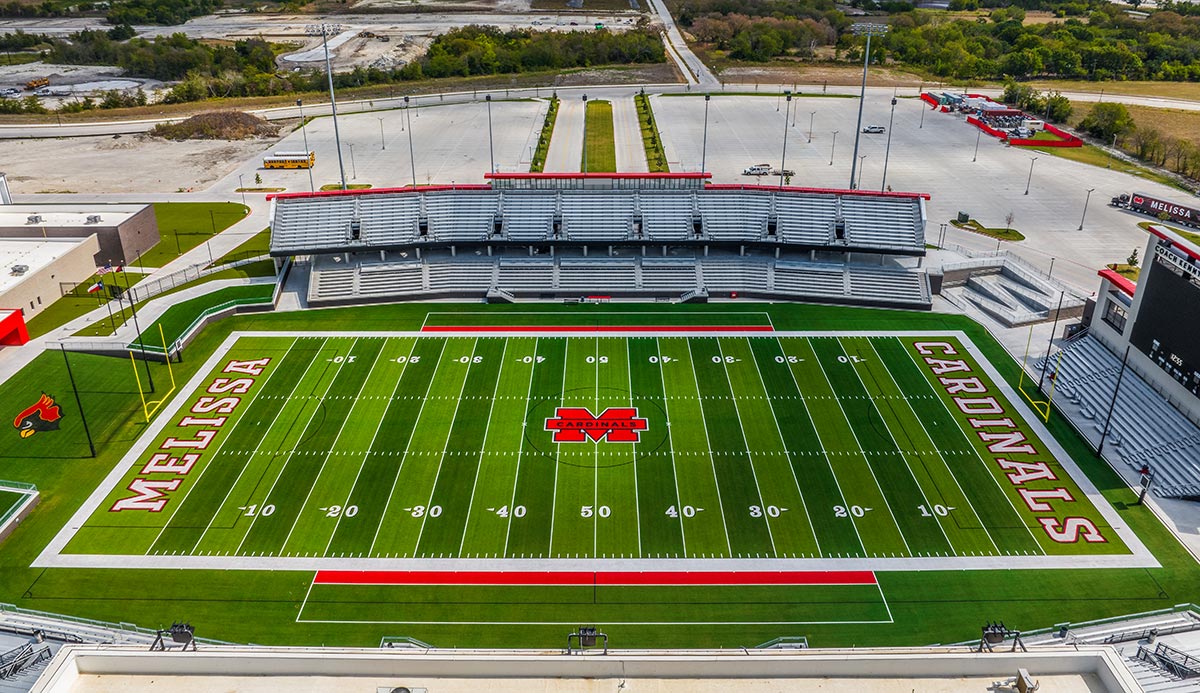 Aerial view of a football field with ‘melissa’ in the left end zone, ‘cardinals’ in the right end zone, and a red cardinal logo at midfield, empty stands nearby.