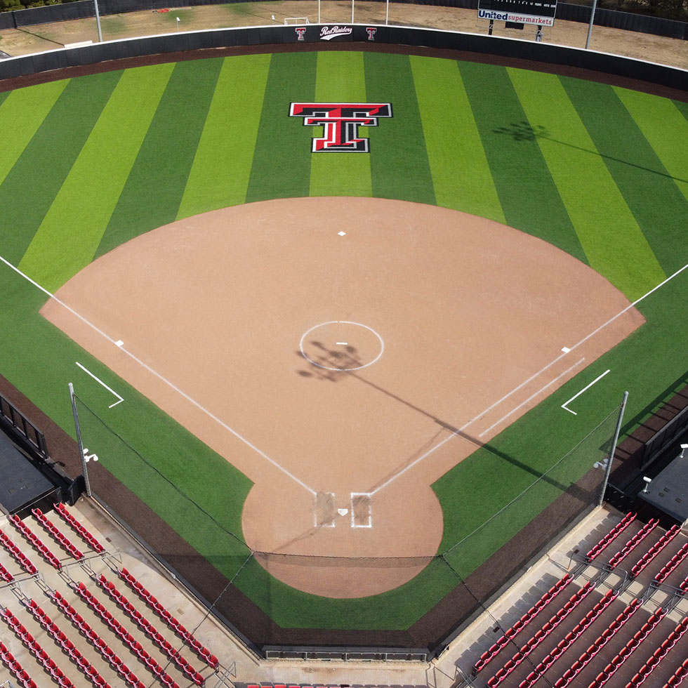 Prestonwood-christian_basketball - Symmetry Sports Construction Aerial view of a baseball stadium with striped green turf, a large tt logo at center field, and red seating surrounding the diamond.