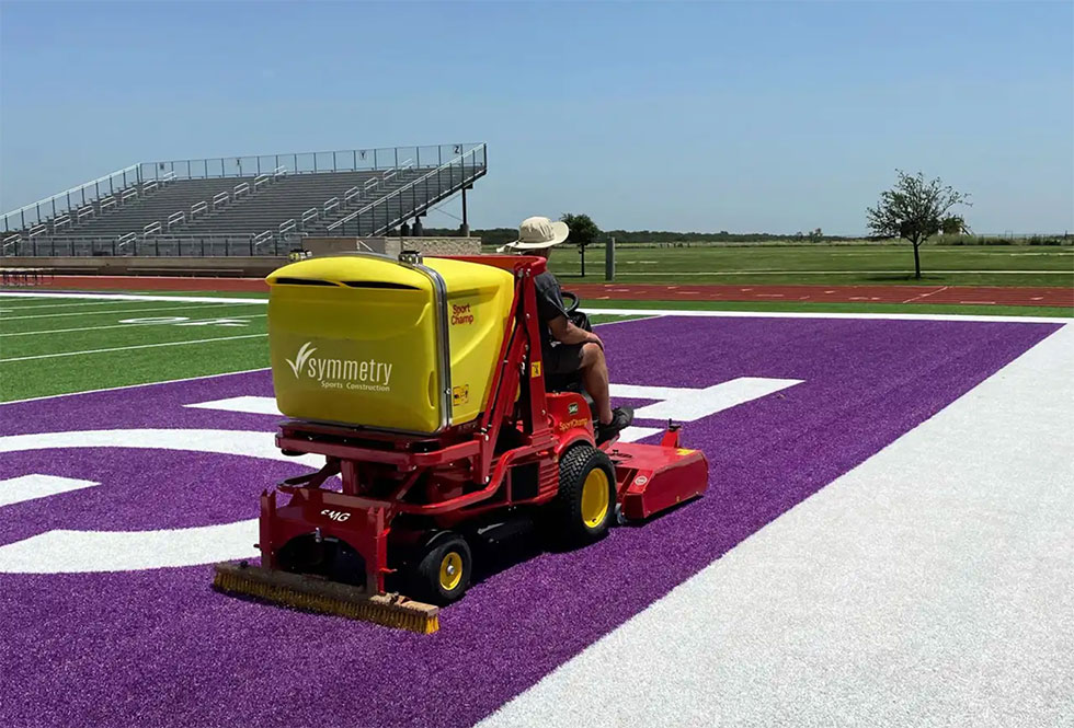 Maintenance rider on a yellow symmetry field-construction machine grooming a purple end zone on an outdoor football field; empty stands in background.